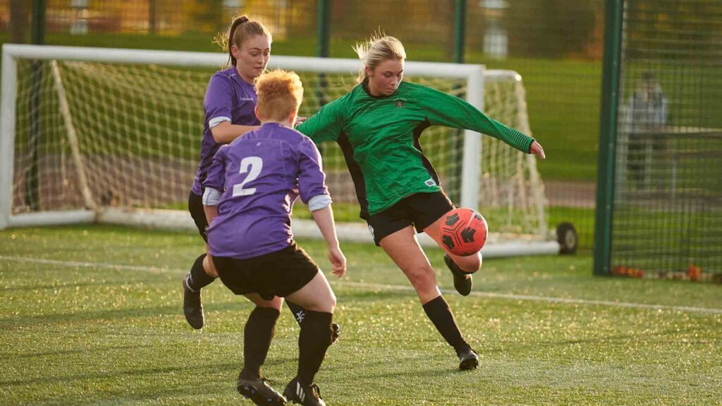 One Edge Hill Women's footballer, wearing green, is mid-kick, with two opponents in purple crowding her.