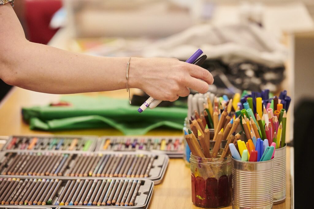 Choosing materials, a hand holding pens reaching to a tub of many mixed coloured pencils