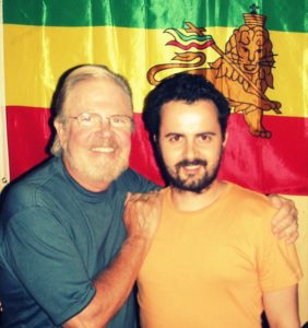 Richard Twine and Tom Regan standing together, smiling at the camera standing in front of brightly coloured flag.