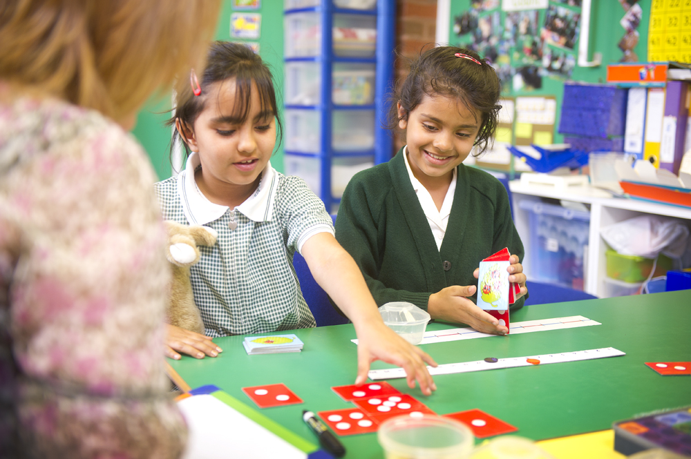 Two children play with cards in a classroom, supervised by their teacher.