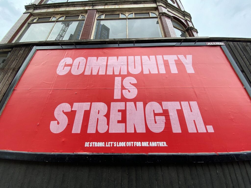 Photo by John Cameron on Unsplash. Community is Strength poster, large red and white public billboard saying 'community is strength'