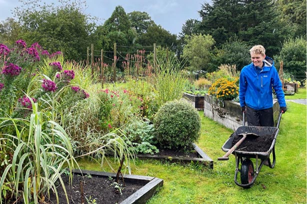 Let's Grow Preston, gardeener carrying wheelbarrow full of soil walking through a garden - pic by LGP