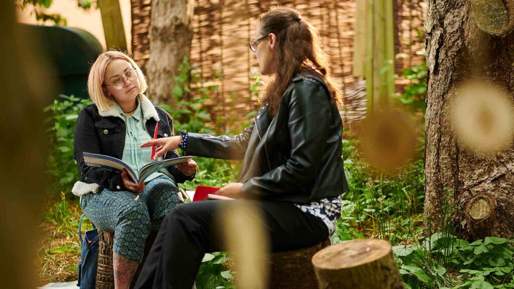 Two women sat outside on tree stumps, one writing in her notepad.