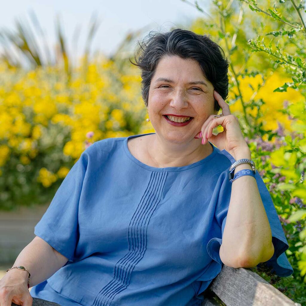 Headshot of Luminita Paraoan. Luminita is wearing a blue blouse and smiling at the camera whilst sitting in the rooftop garden on Edge Hill University's campus.