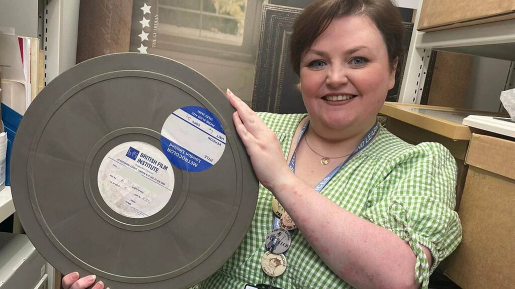 Sophie Smith wearing a green dress and holding an old silver film reel in her hands in between shelves in the archive 