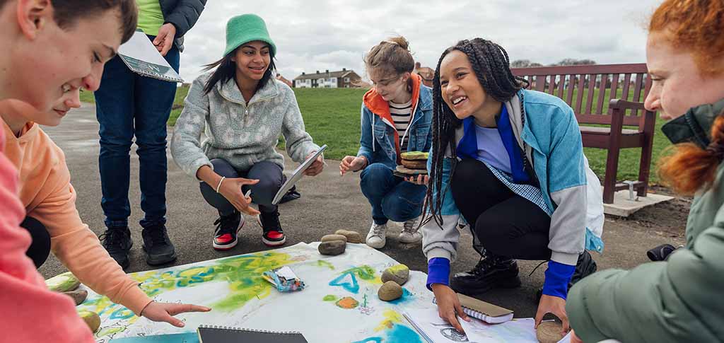 A group of teenage friends wearing casual clothing on an overcast summer day in Whitley Bay, Northeastern England. They are gathered on the promenade by the sea where they use different arts and crafts products to create sustainability artwork to spread a message of environmental conservation for Earth Day.