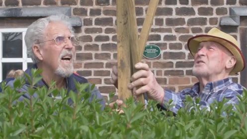 Two older gentlemen holding up a wooden post infront of a brick wall behind a hedge.