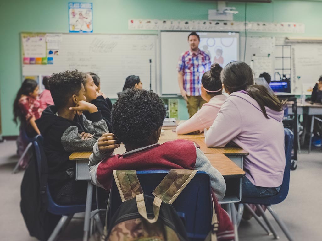 11 kids sitting around tables in a class room being taught by a teacher