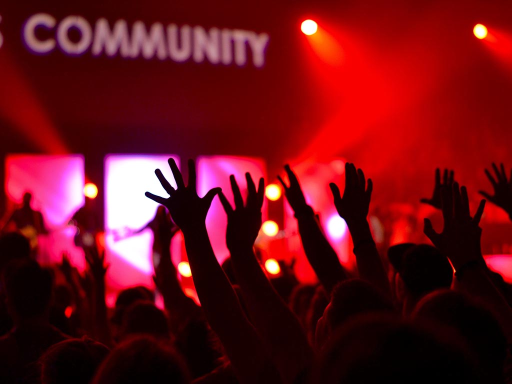 A crow of people at a gig with their hands in the air watching a band, light up with a backdrop of red light