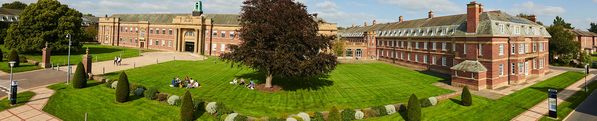 Wide angled image of the main building at Edge Hill University. Students sitting on the grass at the front.